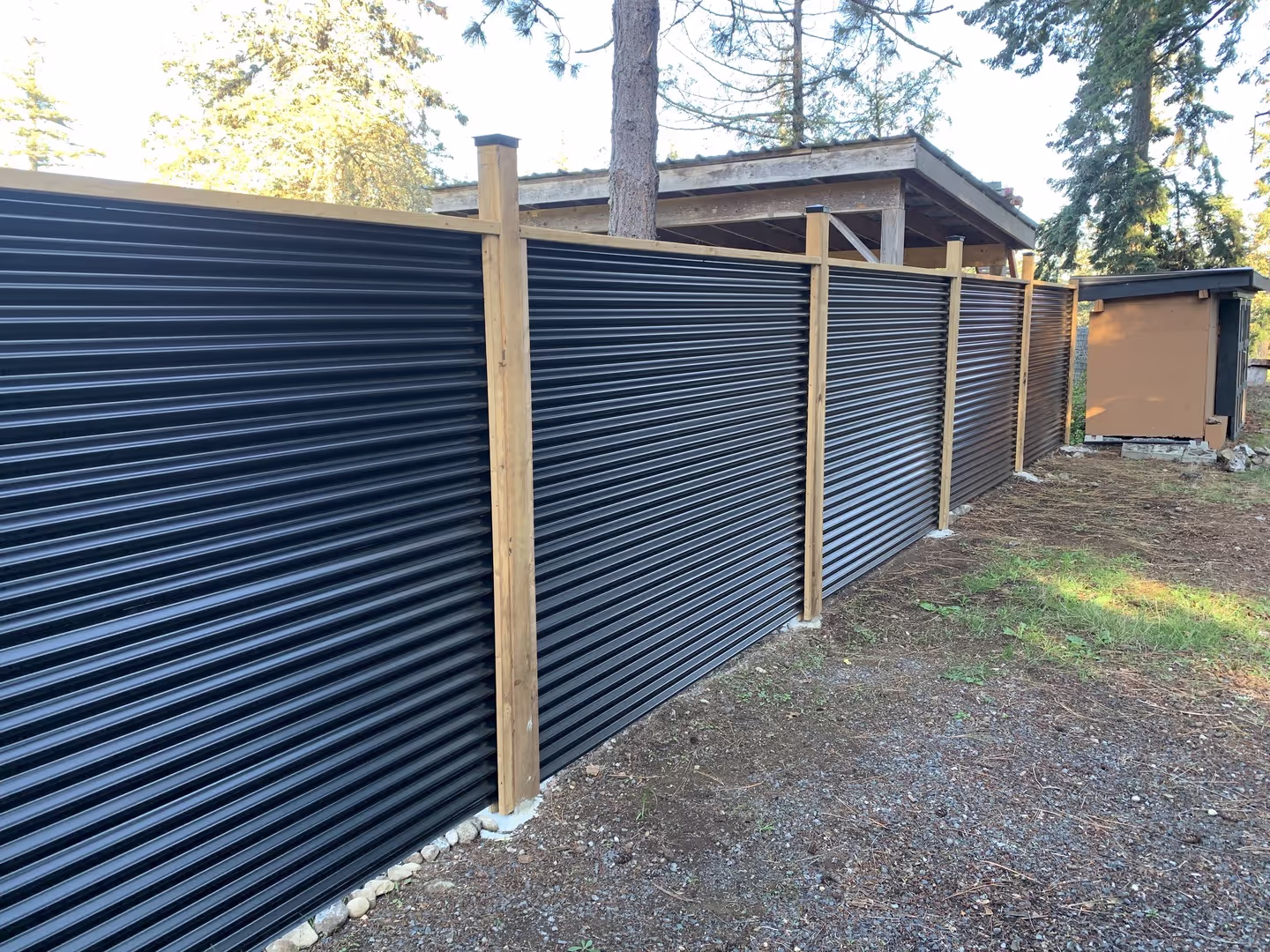 Black corrugated metal privacy fence with wood posts along a backyard with trees.