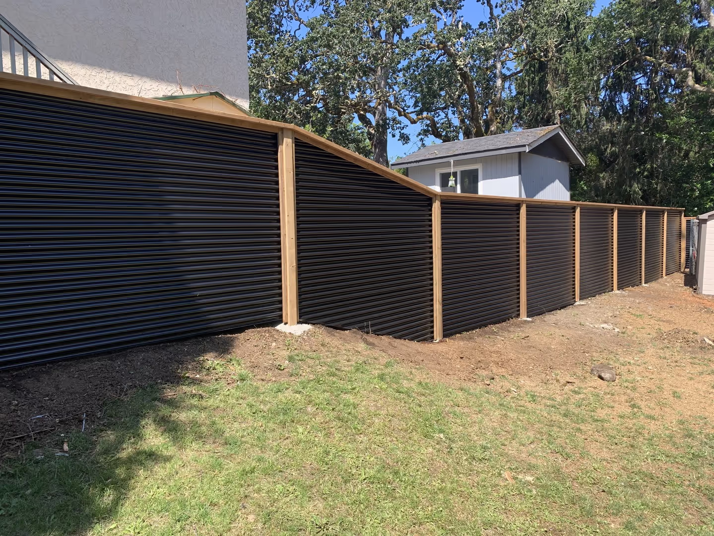 Black horizontal corrugated metal fence with wood posts along a sloped backyard