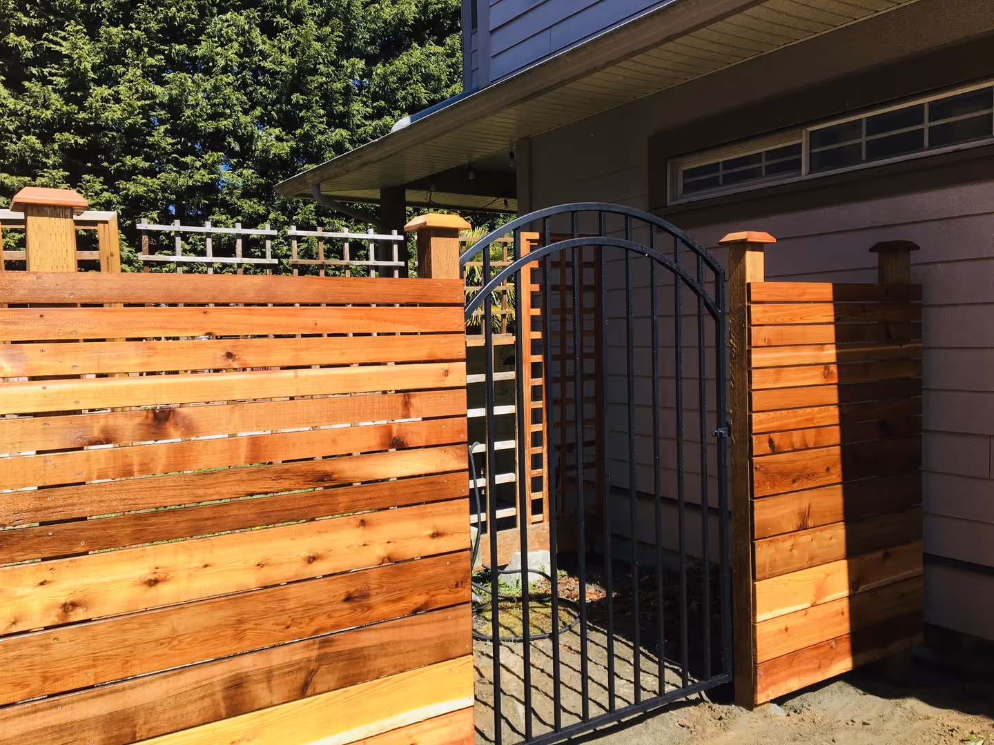 Cedar horizontal-slat privacy fence with arched black metal gate beside a house in sunlight
