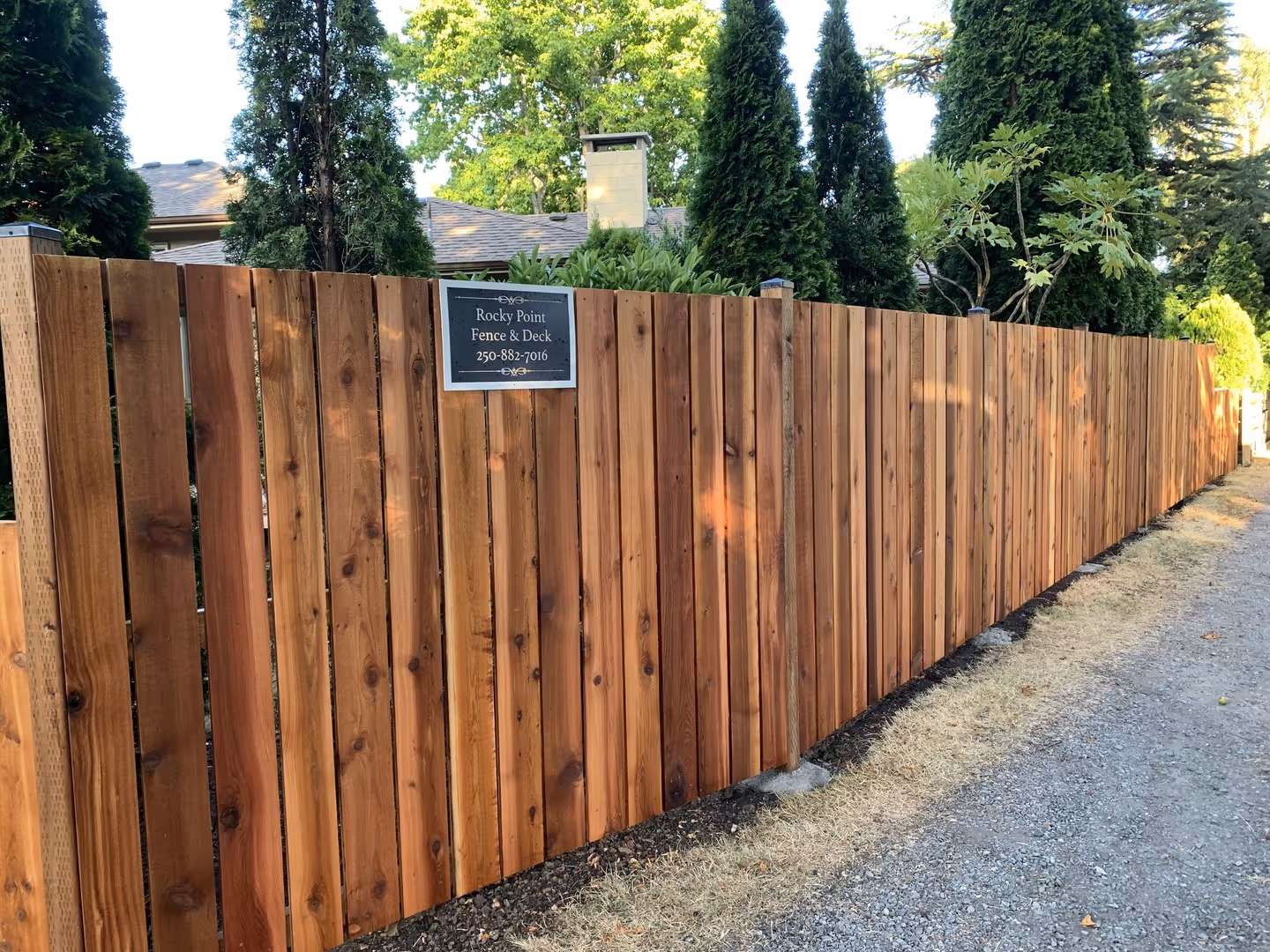 Cedar privacy fence runs along a driveway, with a Rocky Point Fence & Deck sign.