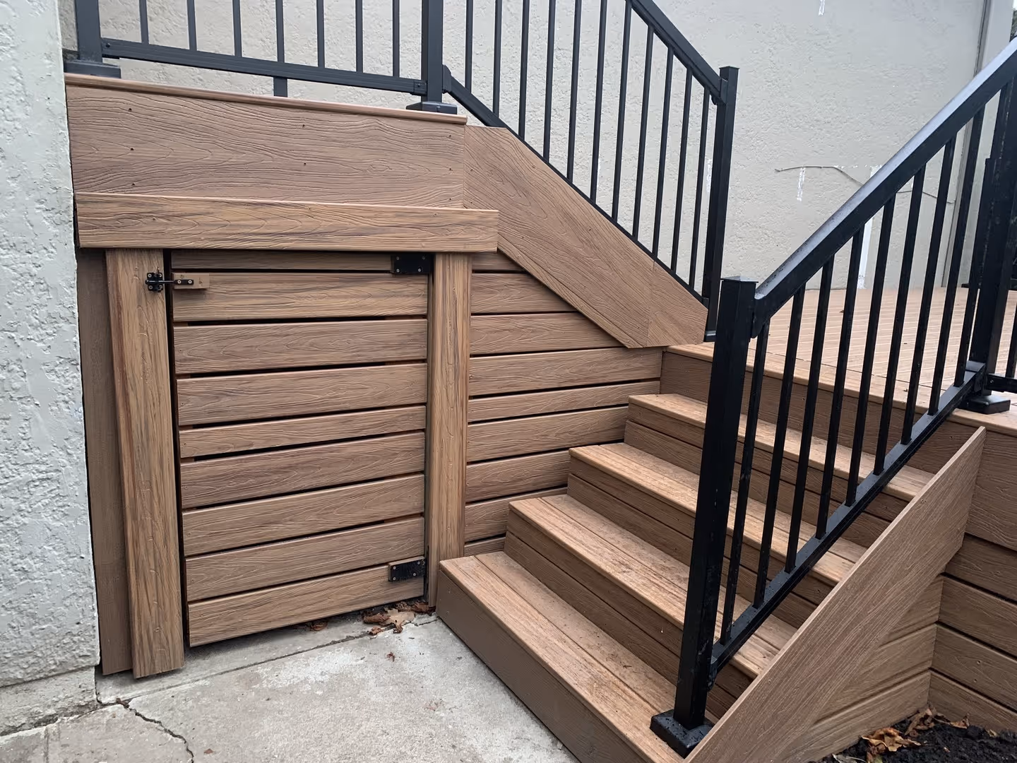 Composite deck stairs with black metal railings and an under-stair storage door beside a concrete wall.