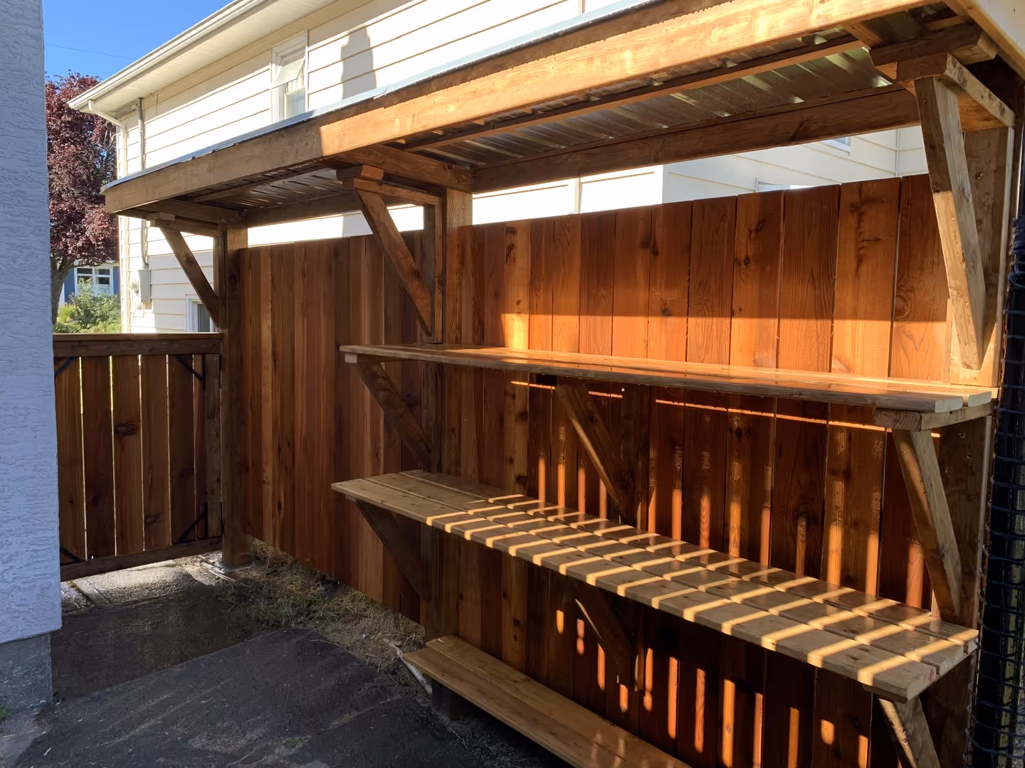 Covered outdoor wooden shelves along a cedar fence near a side yard gate