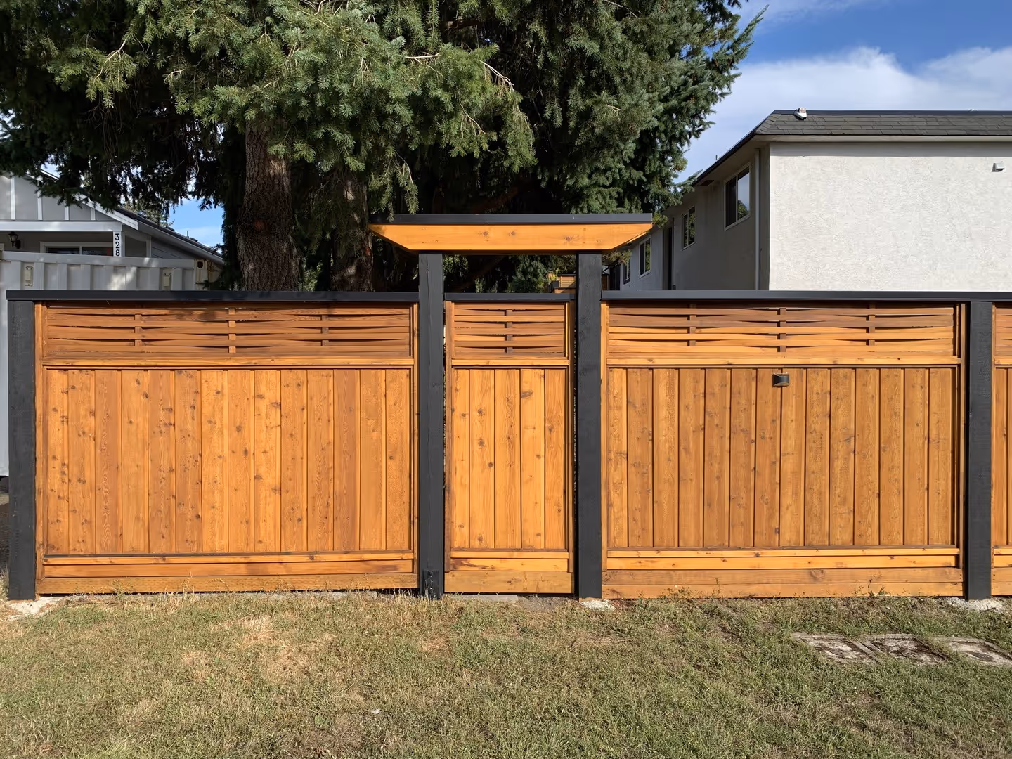 Modern cedar privacy fence with central gate and pergola-style top, black posts, in a residential yard