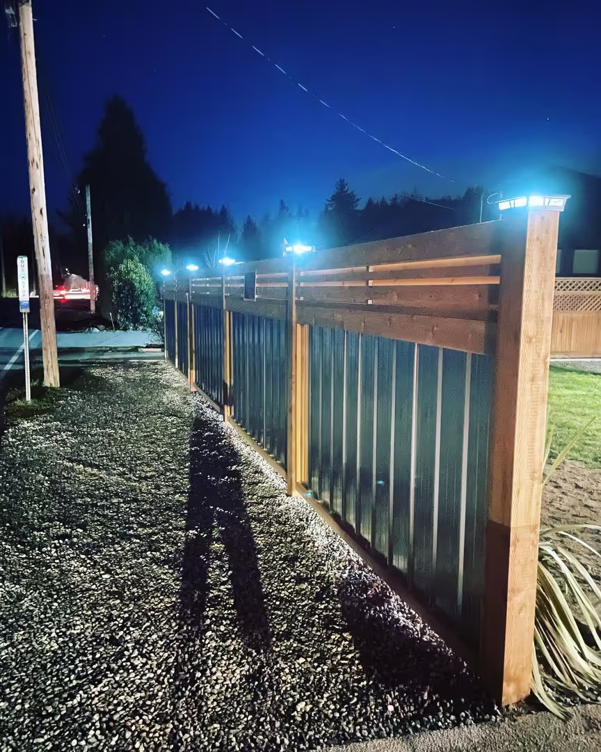 Modern wood-and-metal fence with illuminated post caps at night beside a gravel path.