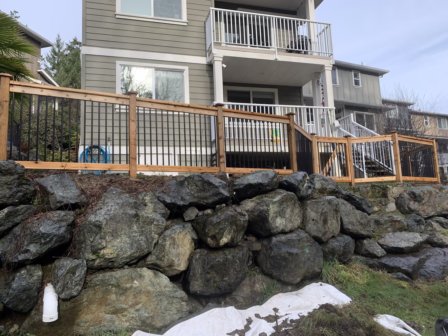 Wood and metal railing fence on a rock retaining wall below a multi-story home.