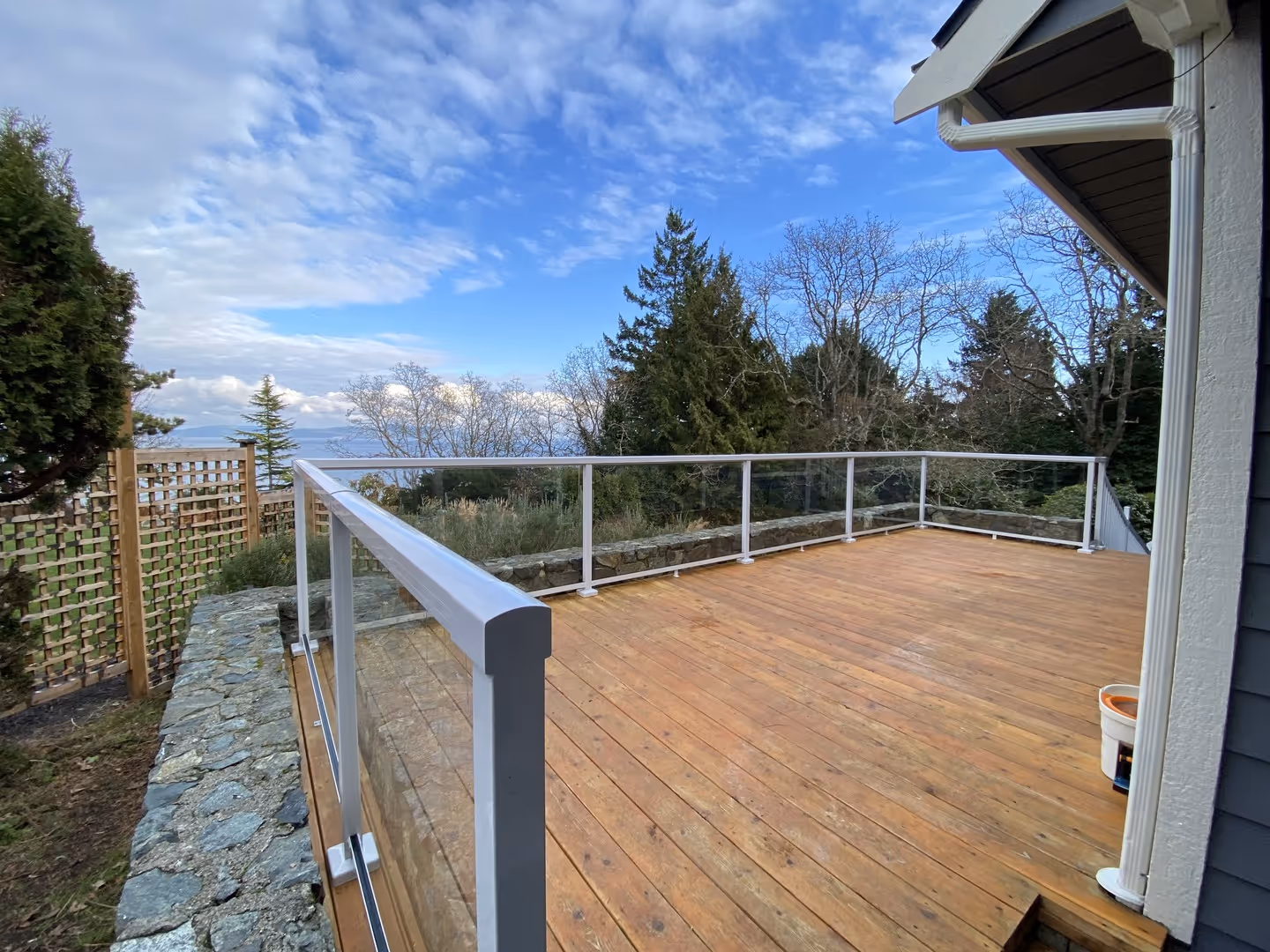 Wood deck with glass railing overlooking trees and water under a blue sky