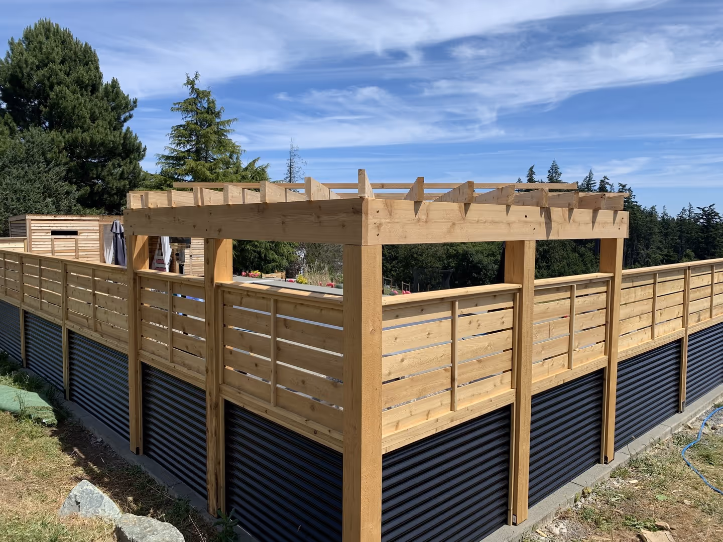 Wooden privacy fence with corrugated metal base and pergola, set against trees and blue sky.