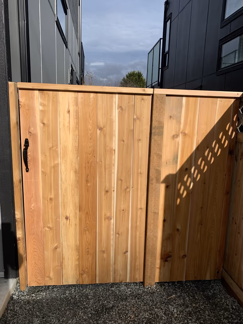 Wooden privacy gate with black handle between modern dark-gray buildings on a gravel path.
